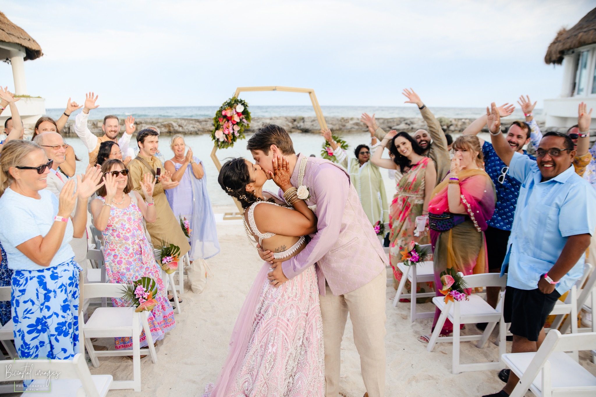 Beautiful beach wedding ceremony in Riviera Maya with couple kissing under geometric floral arch while guests celebrate